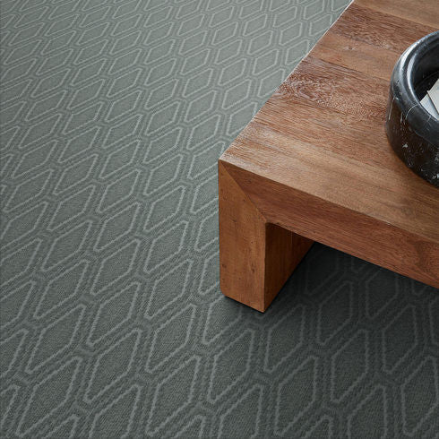 A close-up of a gray patterned carpet with a geometric design, next to the corner of a wooden table and a black ceramic bowl on top.