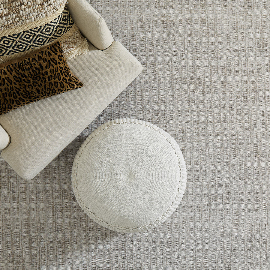 Top-down view of a beige armchair with patterned cushions next to a round, textured white ottoman on a light, textured carpet.