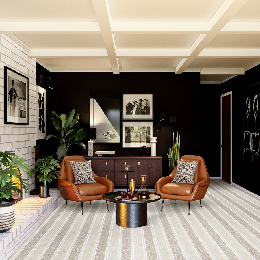 A modern living room with dark walls, two brown leather chairs, a round coffee table, potted plants, and framed art. A striped rug covers the floor and a sideboard holds decorative items against the back wall.