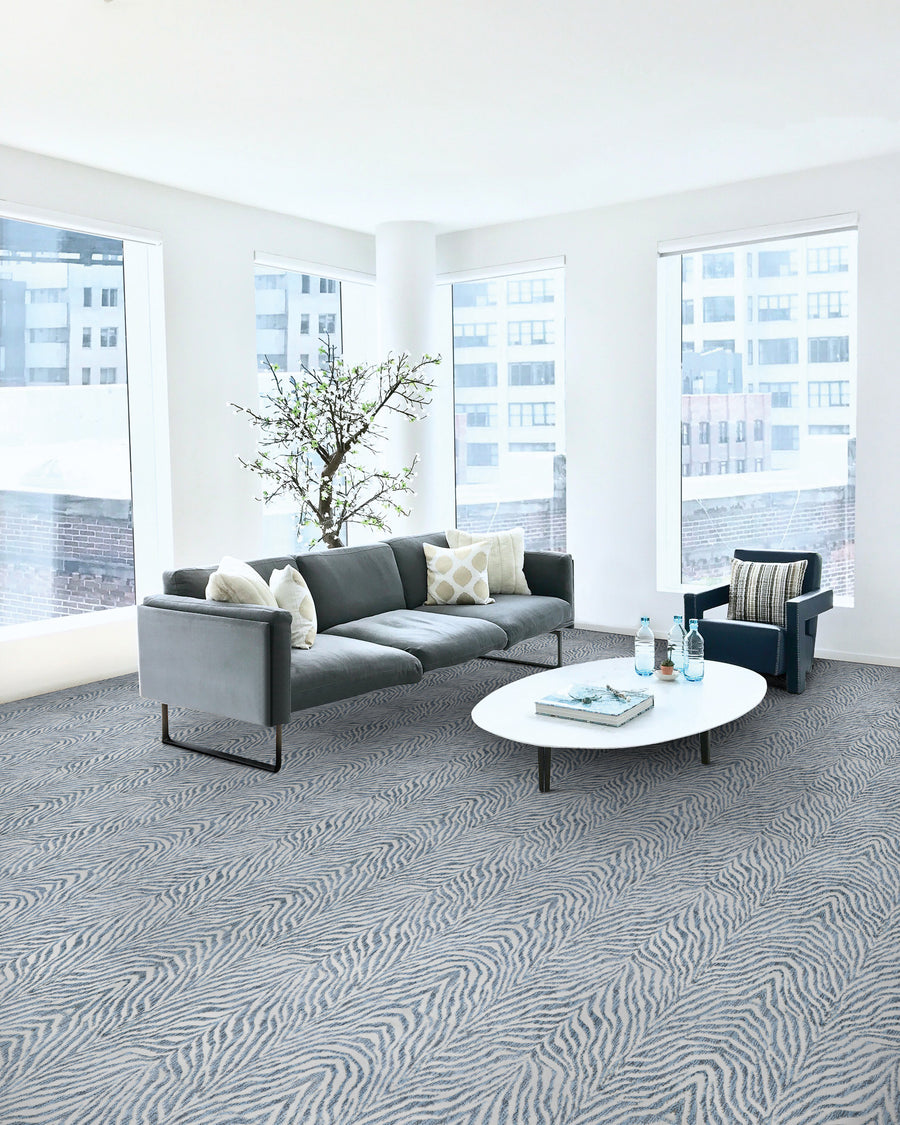 A modern, bright living room with large windows, a gray sofa, a dark blue armchair, a white oval coffee table with drinks, a potted flowering tree, and gray patterned carpet. City buildings are visible outside.