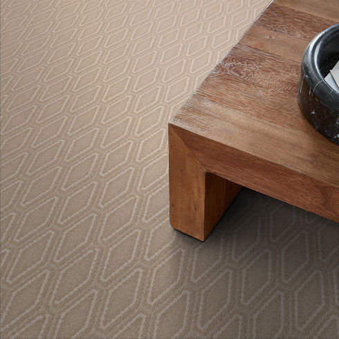 A close-up view of a beige carpet with a textured, geometric diamond pattern next to the corner of a wooden table holding a dark bowl.