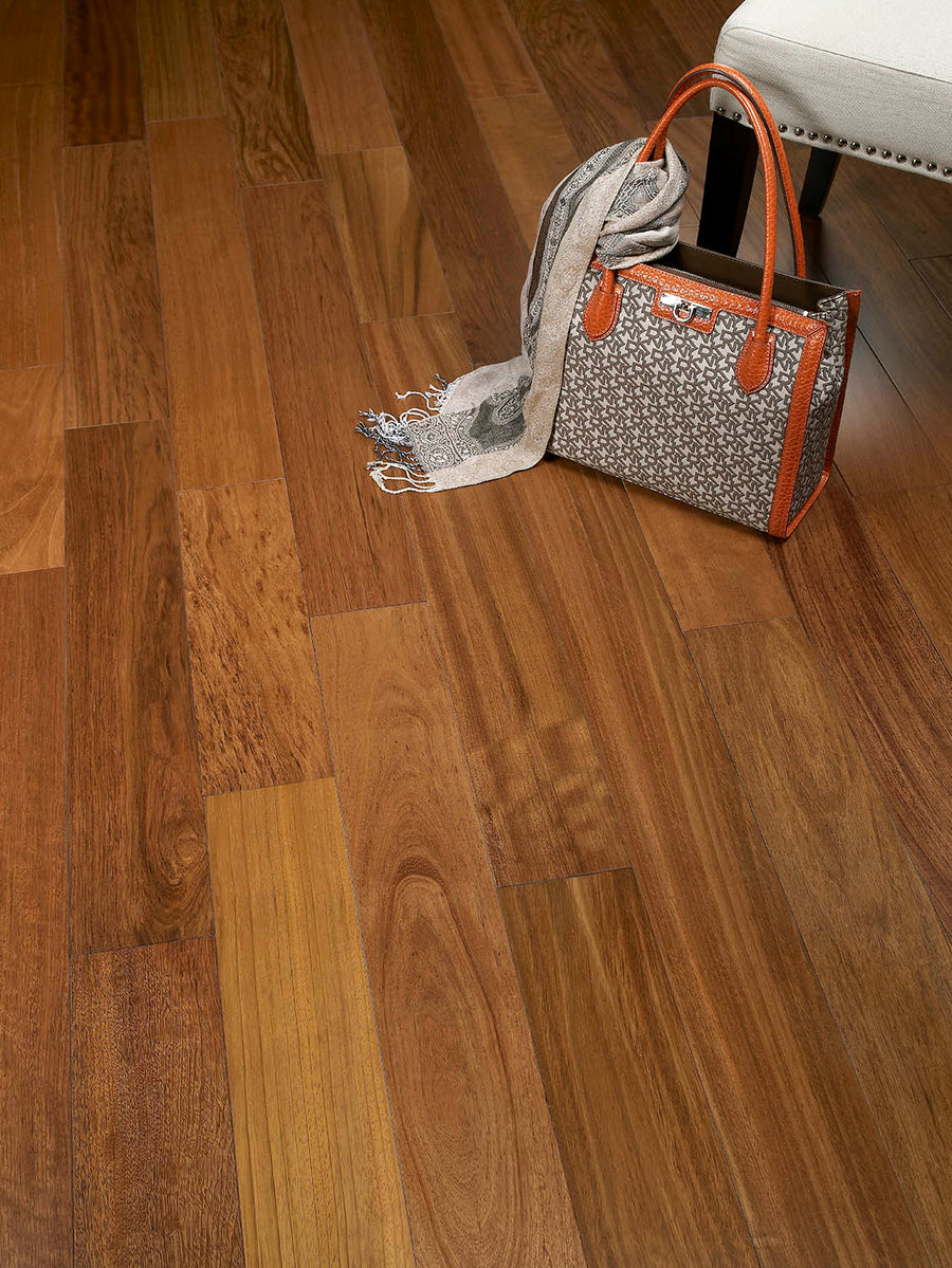 A brown patterned handbag with orange trim and a gray scarf rest on a light-colored upholstered chair, set on a polished wooden floor with natural grain patterns.
