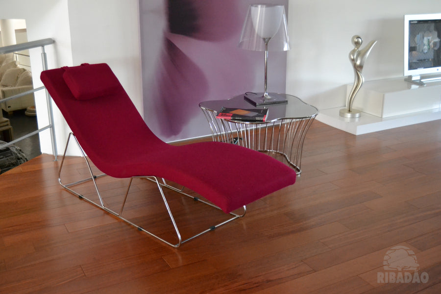 A modern living room with a red chaise lounge, a metallic side table holding books and a lamp, wooden flooring, abstract wall art, and a silver decorative sculpture near a TV.