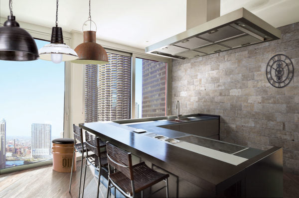 Modern kitchen with a large island, metal stools, industrial pendant lights, and a brick accent wall. Floor-to-ceiling windows offer a city skyline view with tall buildings. A wall clock hangs on the right.