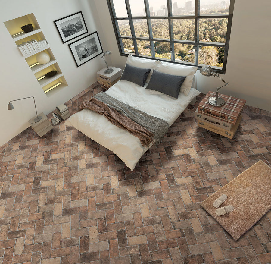 A cozy bedroom with a large window, a bed with gray and white bedding, nightstands, a lamp, wall shelves, framed photos, a plaid ottoman, a rug with slippers, and brown herringbone tile flooring.