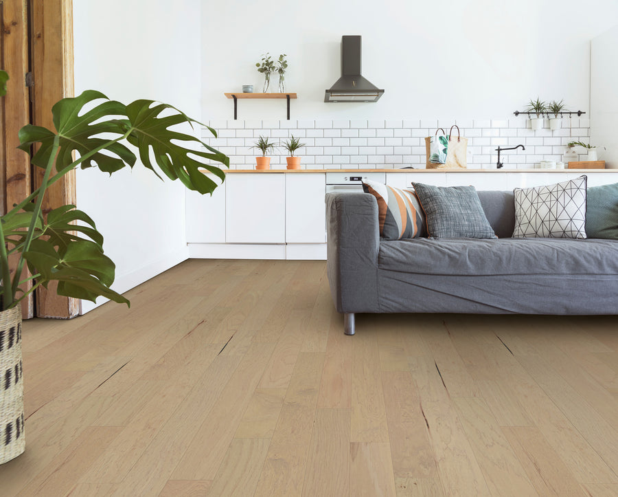 Modern living room featuring elegant Deepwood Trail 5″ Hickory Oak Hardwood flooring, a gray sofa adorned with patterned pillows, a potted plant in the foreground, and a white kitchen with open shelves, greenery, and a black range hood in the background.