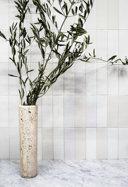 A tall, textured beige vase with long, leafy green branches sits on a marble countertop in front of a white tiled wall.