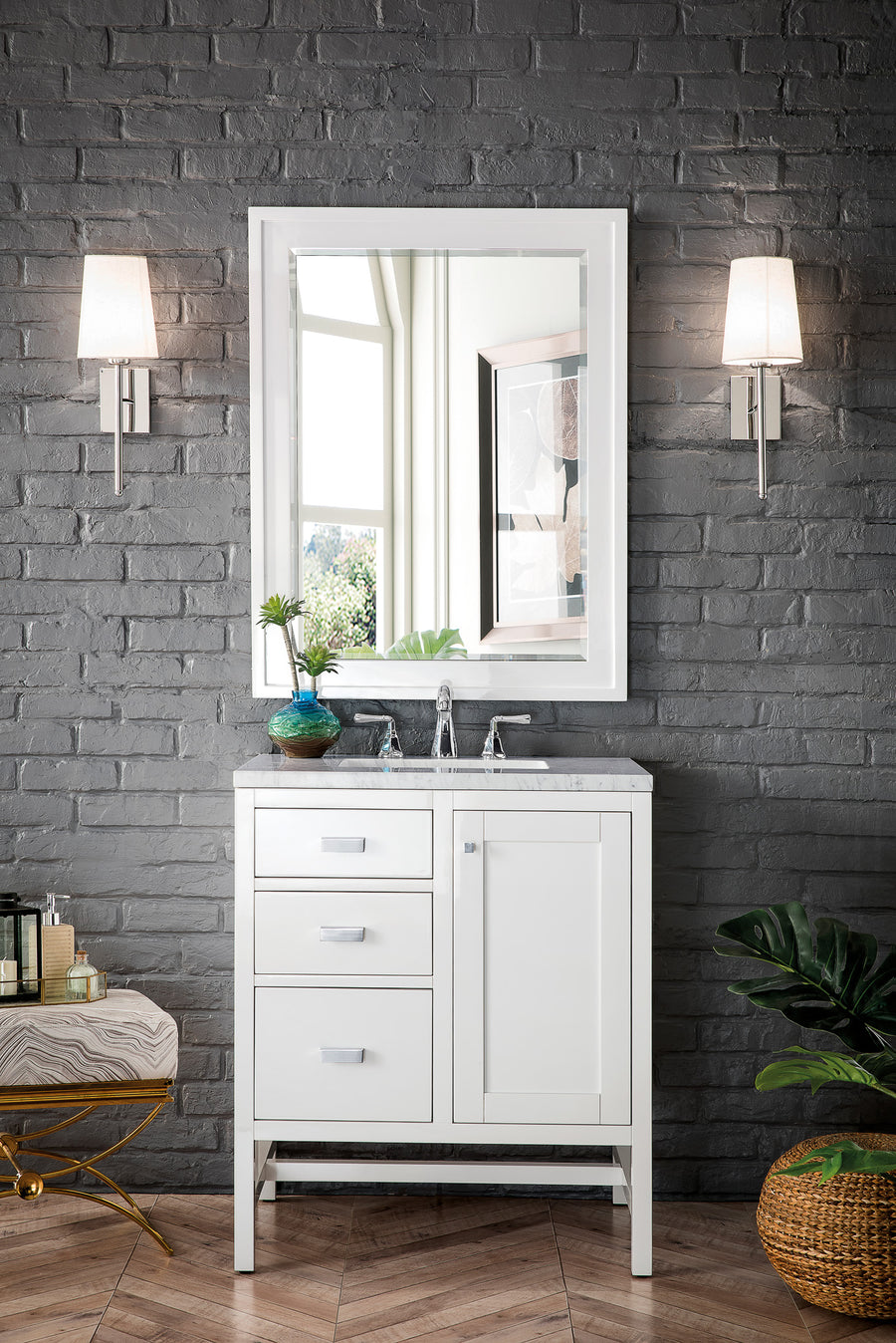 A modern bathroom vanity with a white cabinet, marble countertop, and a rectangular mirror, set against a dark gray brick wall. Two wall sconces with white shades are on each side of the mirror, and plants sit nearby.