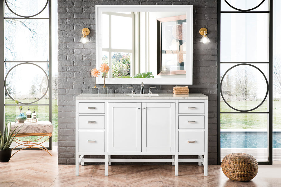 A modern bathroom with a white double-sink vanity, a large mirror, two wall sconces, and gray brick walls. Large windows showcase a green outdoor view, and a woven ottoman sits on a herringbone wood floor.