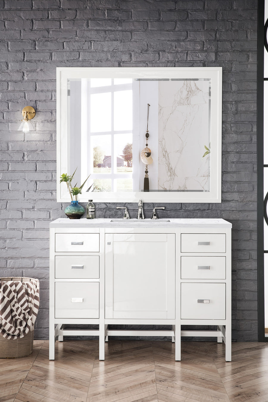 A modern bathroom vanity with a large white-framed mirror, white sink cabinet with drawers, and silver faucet against a gray brick wall. A round woven basket sits to the left, and a wall sconce is mounted above.