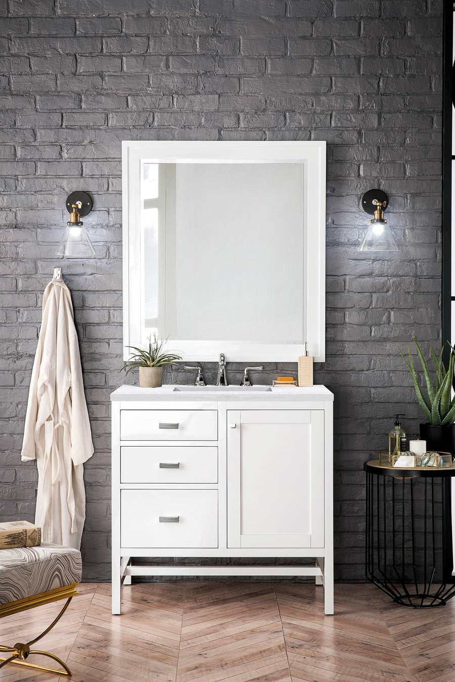 A modern bathroom with a gray brick wall, a white vanity with drawers, a large mirror, two wall sconces, potted plants, a hanging robe, and a small side table on a herringbone wood floor.