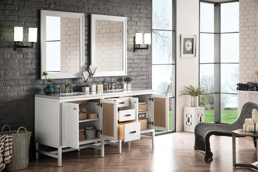 A modern bathroom with a white double-sink vanity, two mirrors, open drawers and cabinets, wicker baskets, wall lights, and large windows overlooking greenery and a lake. A woven chair sits to the right.