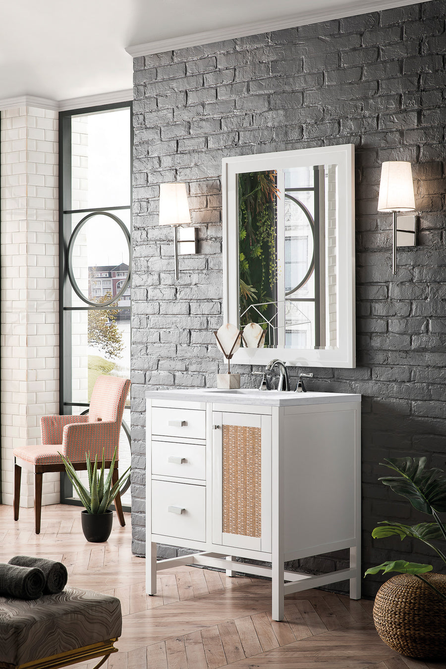 A modern bathroom with a white vanity, wicker cabinet detail, wall mirror, and two sconces on a gray brick wall. Large windows and indoor plants create a bright, airy atmosphere with wood flooring.