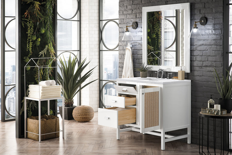 A modern bathroom with a white vanity, open drawers, a mirror, indoor plants, stacked towels, and large windows showing a city view. The walls are gray brick, and greenery decorates the space.