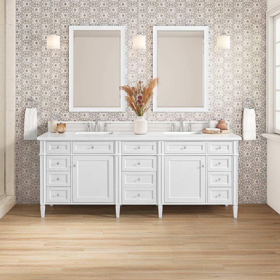 The Brittany 84" Double Bowl Vanity features white cabinets made from Birch and White Poplar, twin mirrors, pendant lights above each sink, patterned wallpaper backdrop, and a central vase of dried flowers for added charm.