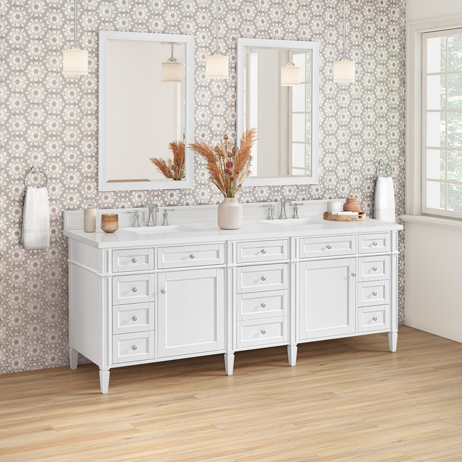 A bright bathroom with a double-sink white vanity, two mirrors, pendant lights, patterned wallpaper, and wooden floor. Decorative items, such as vases and towels, are neatly arranged on the countertop.