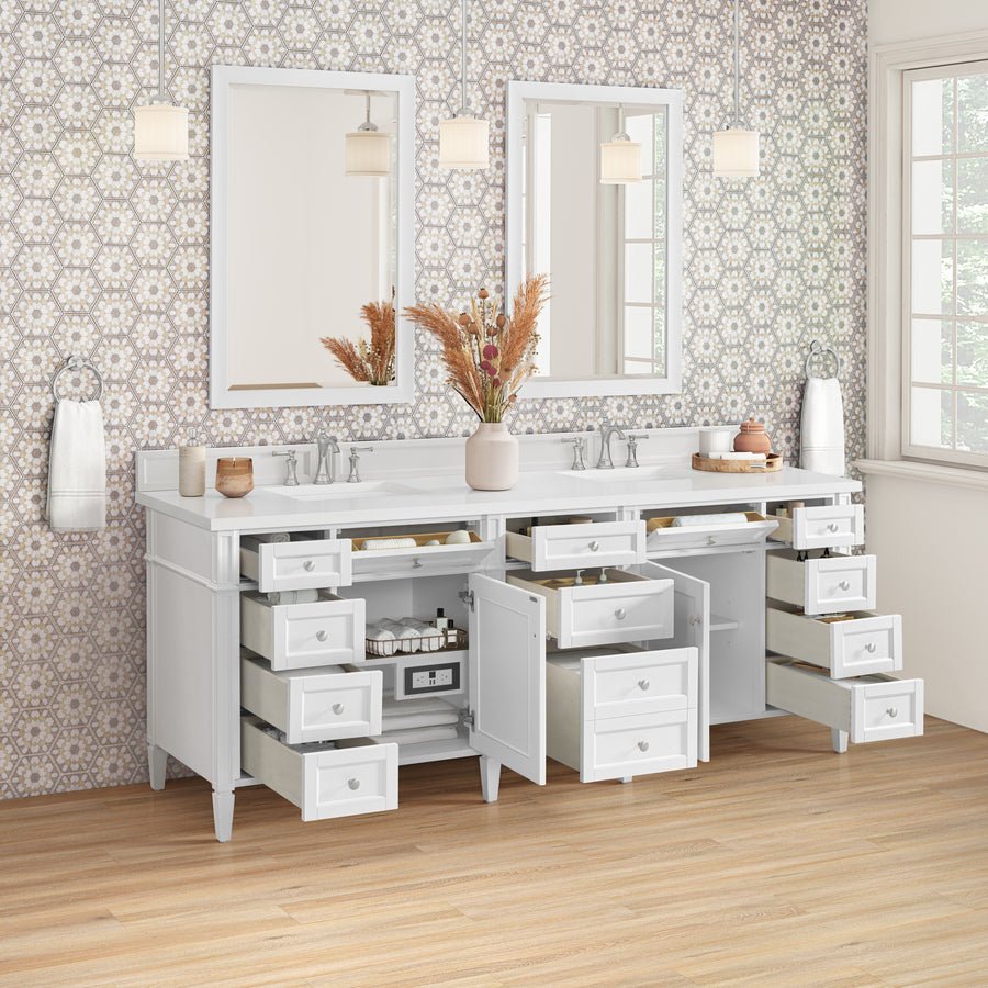 A double-sink white bathroom vanity with all drawers and cabinets open, revealing storage space and plumbing. Two mirrors, three pendant lights, and a vase with dried plants sit on a white countertop against patterned wallpaper.