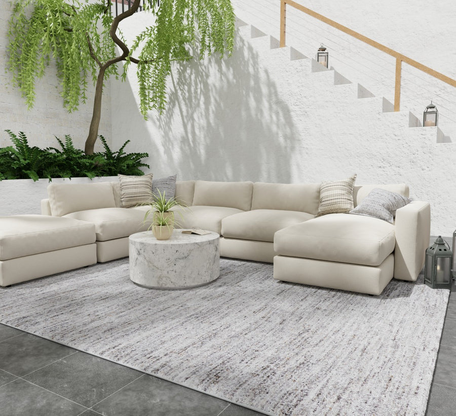 A modern outdoor seating area with a large cream-colored sectional sofa, round marble coffee table, and neutral rug. Green plants and a white stairway with lanterns add a serene touch to the bright, airy space.