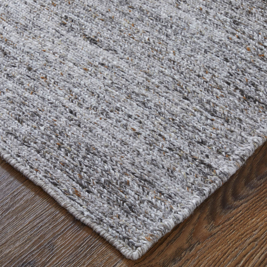 Close-up of the corner of a textured, woven gray rug with flecks of brown, beige, and black, placed on a dark wooden floor.