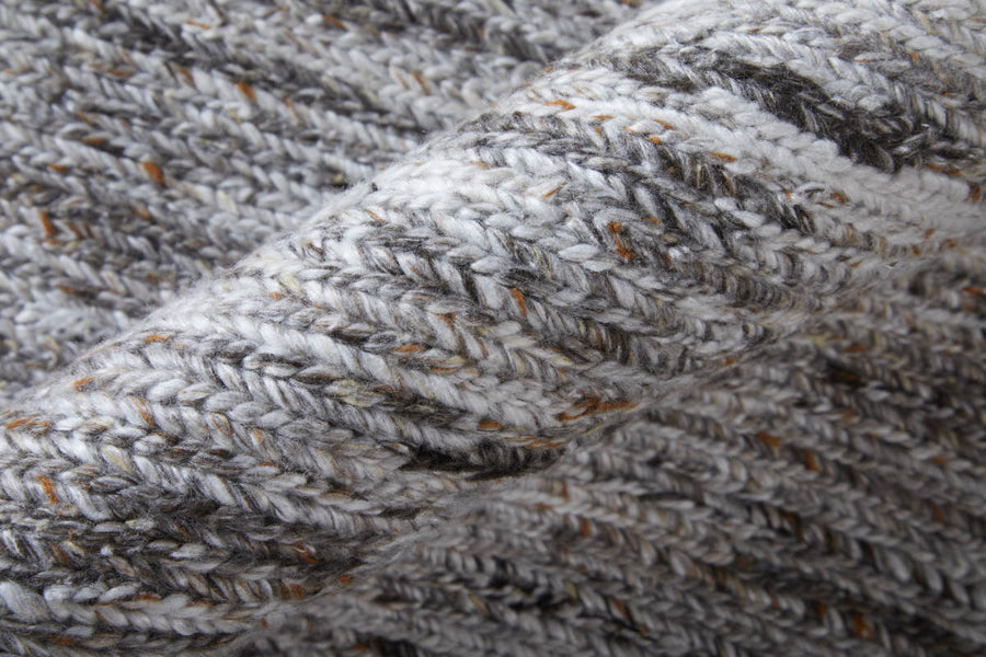 Close-up of a folded, knitted wool fabric in shades of grey and white, with visible textured stitches and flecks of brown yarn woven throughout.