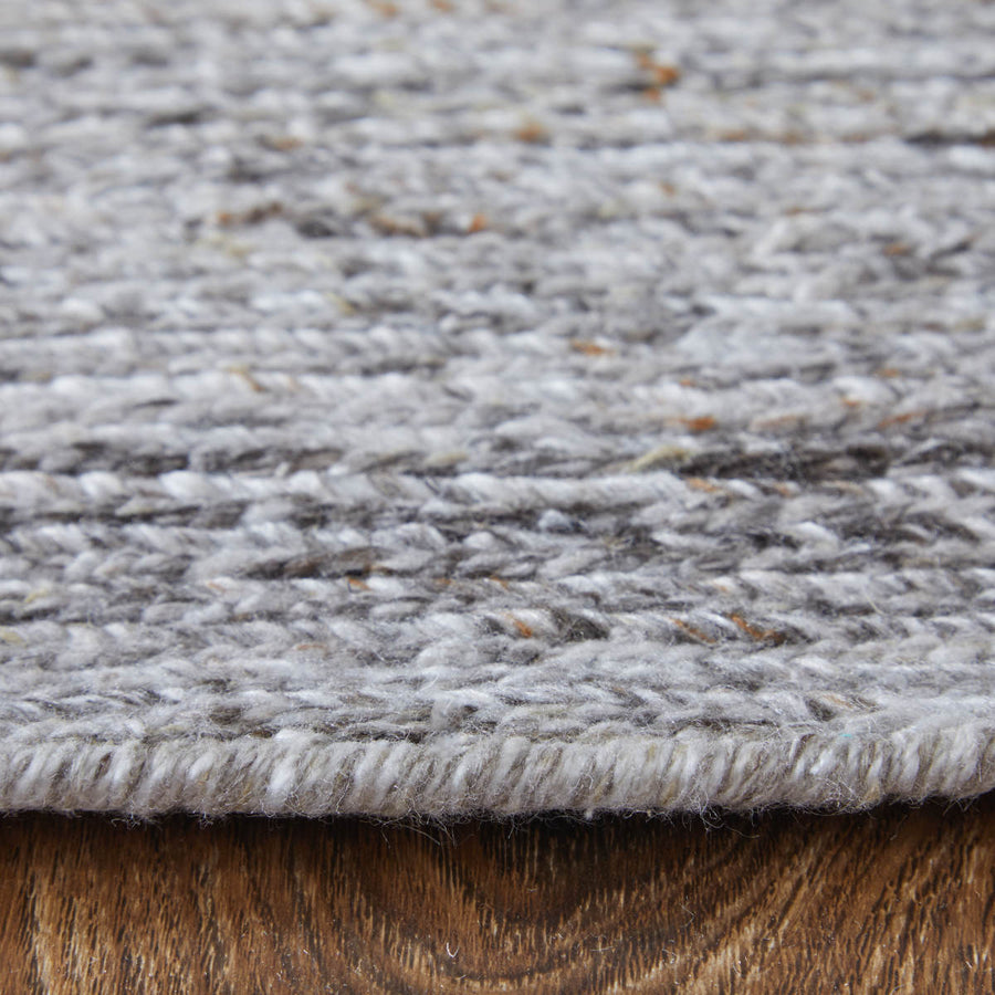 Close-up side view of a gray woven rug with a thick texture, lying on a brown wooden floor. The image highlights the rugs weave and thickness.