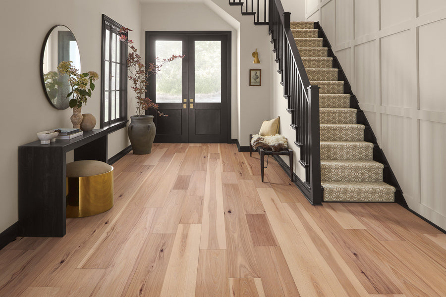 A modern entryway with light wood floors, a black console table with a round stool, a large floor vase with branches, a round mirror, and a carpeted staircase with black railing next to double front doors.
