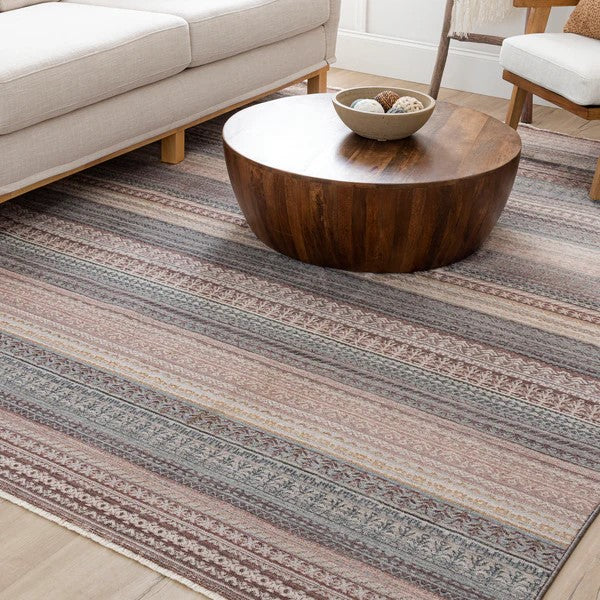 A round wooden coffee table sits on a striped area rug with muted earth tones in a modern living room, next to a light-colored sofa and a decorative bowl on the table.