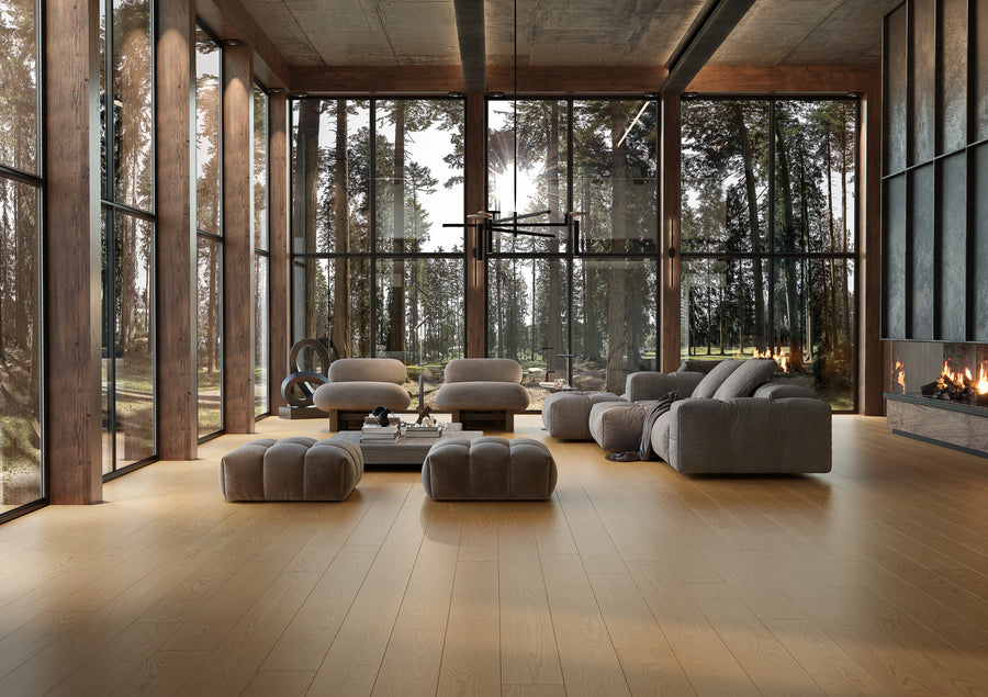 Modern living room with large floor-to-ceiling windows, gray sofas and ottomans, wooden floor, and a fireplace. Lush forest scenery is visible outside, filling the space with natural light.