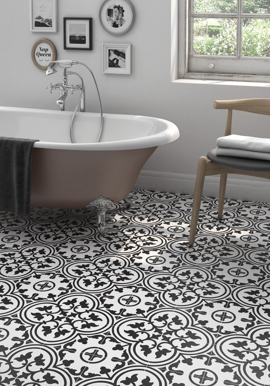 A stylish bathroom featuring Casablanca 8x8 Glazed Ceramic Patterned Tile, a white clawfoot tub draped with a gray towel, European wall art, a wooden chair holding folded towels, and natural light streaming through the window.