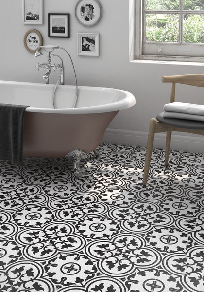 A stylish bathroom featuring Casablanca 8x8 Glazed Ceramic Patterned Tile, a white clawfoot tub draped with a gray towel, European wall art, a wooden chair holding folded towels, and natural light streaming through the window.