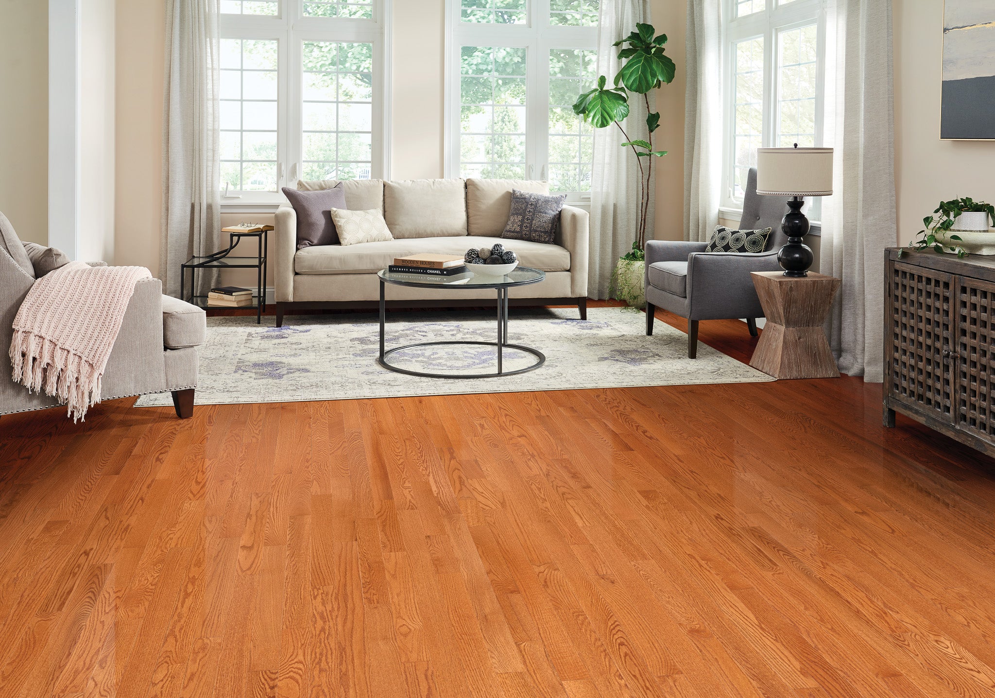 
		                            	Bright living room with large windows, wooden floors, a beige sofa, gray armchair, round glass coffee table, area rug, potted plant, and modern decor including a lamp and console table.
		                            