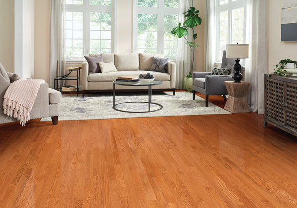 Bright living room with large windows, wooden floors, a beige sofa, gray armchair, round glass coffee table, area rug, potted plant, and modern decor including a lamp and console table.