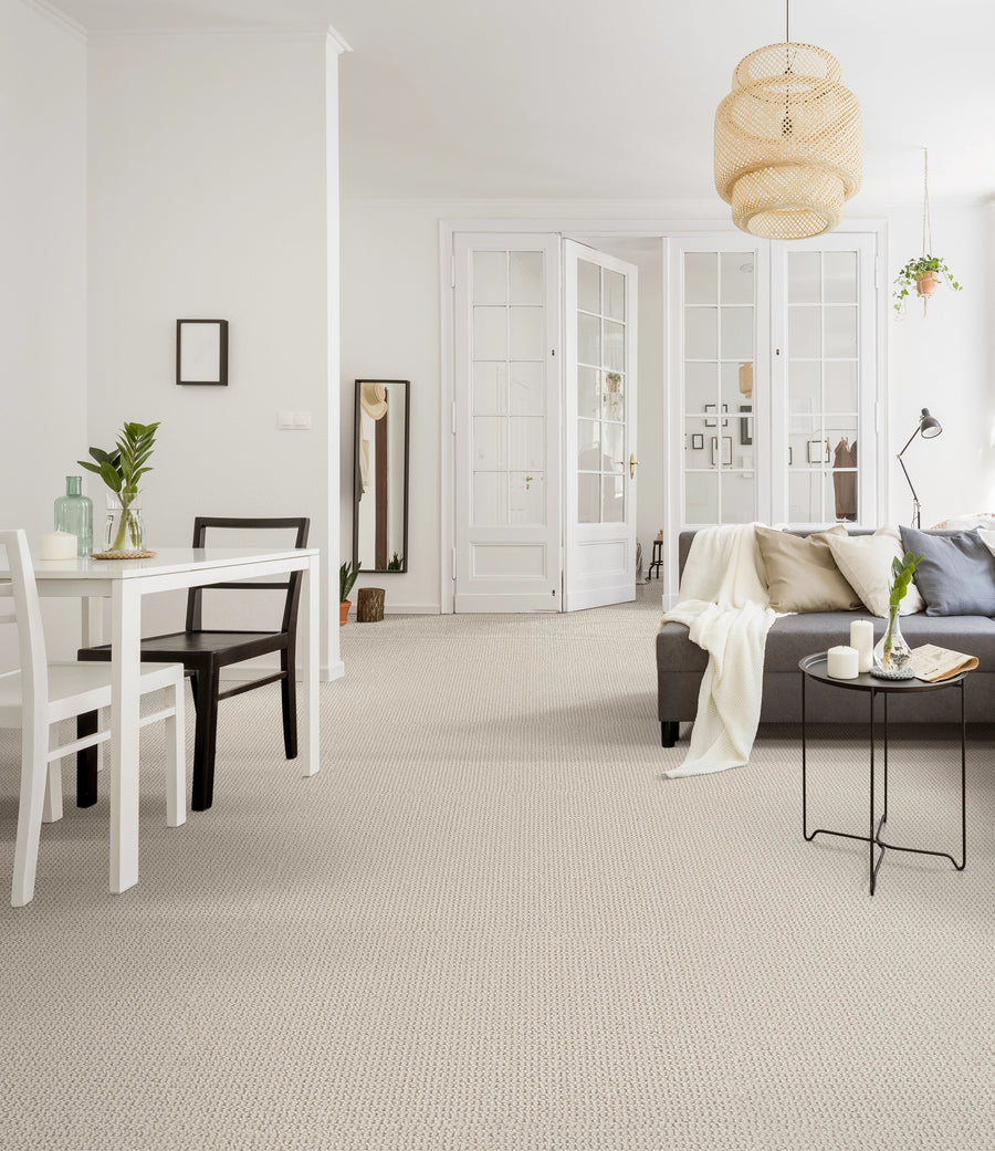 Bright, modern living and dining area with white walls, light carpet, a gray sectional sofa with pillows, a small black side table, a dining table with mismatched chairs, and glass double doors in the background.