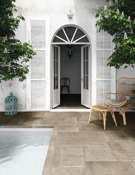 A serene patio with stone tiles, a pool, wicker chair and small table, white French doors with arched window, and leafy green trees beside a white stucco wall.