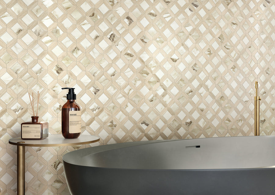 A modern bathroom showcases a beige and white geometric wall with Corsica Lux Mosaic Porcelain Tile, a gray freestanding tub, gold faucet, and a small round side table holding two brown bottles and a reed diffuser.