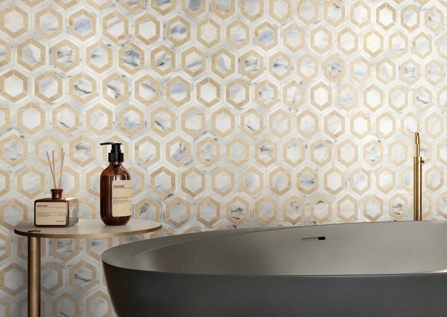 A modern bathroom with a geometric gold and white Corsica Evo Hex Mosaic Porcelain Tile wall, a sleek gray bathtub, and a round side table topped with brown bottles and a reed diffuser.