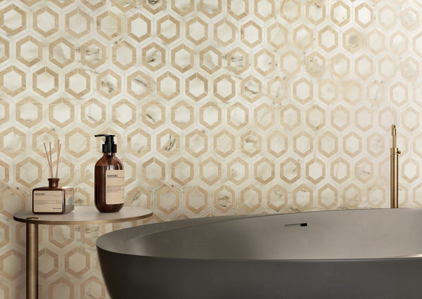 A modern bathroom featuring a wall clad in Corsica Evo Hex Mosaic Porcelain Tile, a freestanding matte gray bathtub, and a round side table with brown bottles and a reed diffuser.