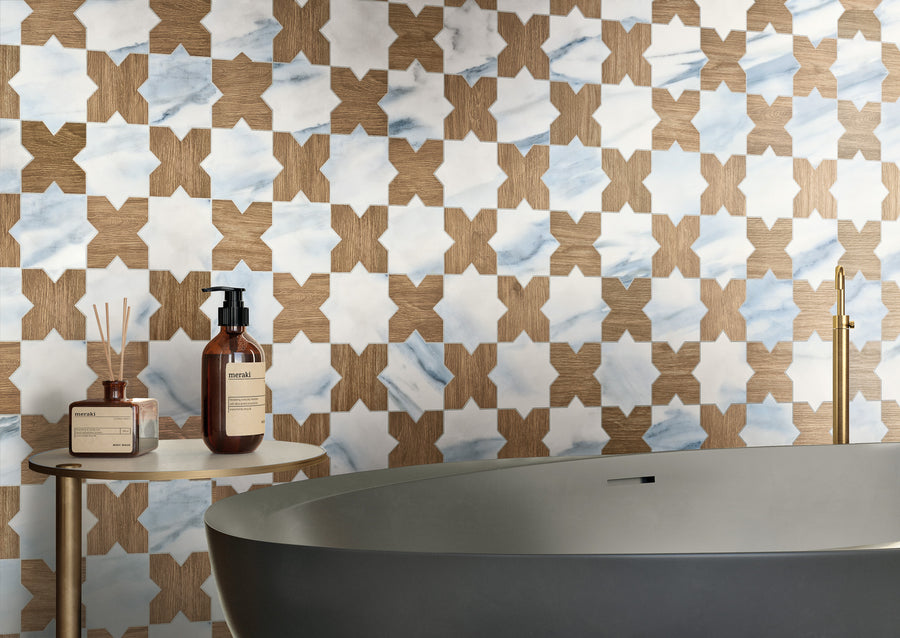A modern bathroom features a patterned wall with star-shaped tiles in wood and white-blue marble. A gray bathtub sits beside a small round table holding bottles and a reed diffuser.