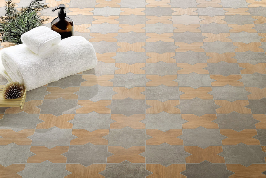 A close-up of a patterned tile floor with interlocking star shapes in beige and gray. On the floor are two rolled white towels, a soap dispenser, a brush, and a small branch, suggesting a spa setting.