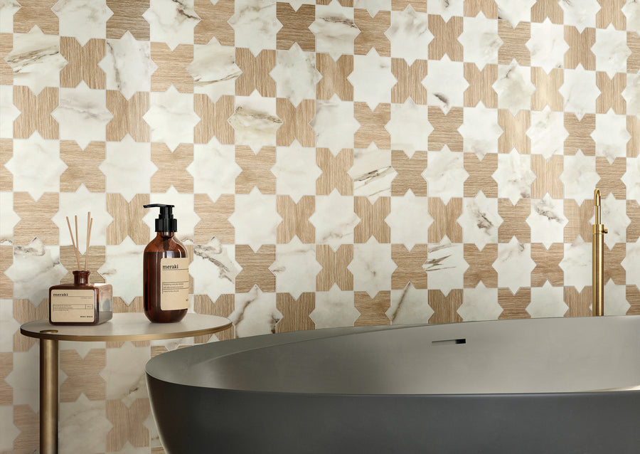 A modern bathroom with a freestanding gray tub, a small round side table holding brown bottles, and a wall featuring a geometric beige and white tile pattern.