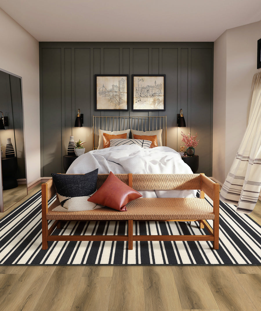 A modern bedroom with a cozy bed, decorative pillows, and a wooden bench at the foot. The room features a dark accent wall, framed art, striped rug, and hardwood floors with soft natural lighting.