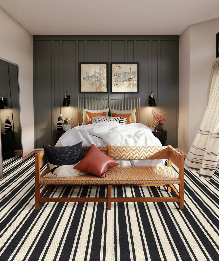 Modern bedroom with a double bed, two framed artworks above the headboard, a wooden bench with cushions at the foot of the bed, striped black and white rug, and decorative lamps on bedside tables.