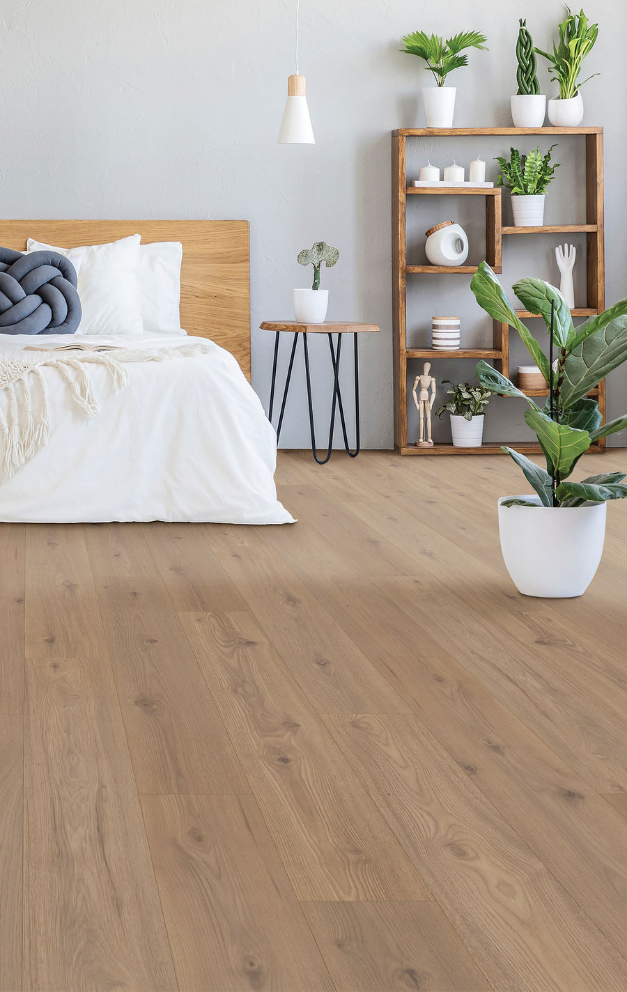 A modern bedroom featuring Freywood Springs 8 11/32" Waterproof Laminate flooring, a wooden bed with white bedding, a round side table, potted plants, and a wooden shelf with books and decor against a light gray wall.