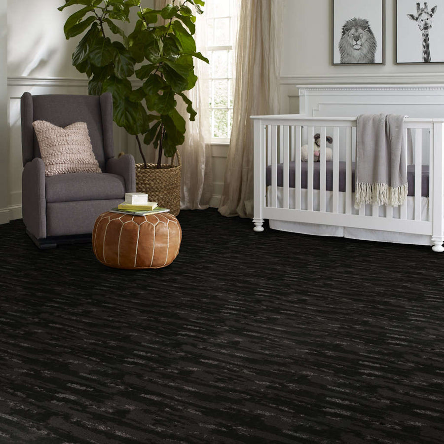A cozy nursery with a white crib, gray armchair with a pink pillow, tan pouf, large leafy plant, and animal artwork on the wall. The room has dark patterned carpet and natural light from a window.