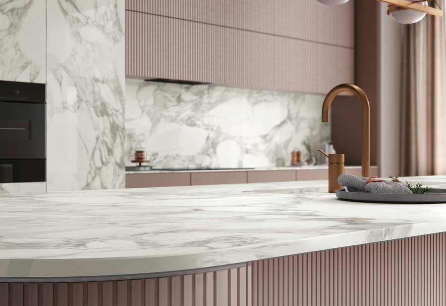 Modern kitchen with a large white marble countertop, matching marble backsplash, matte brown cabinets, a copper faucet, and a tray with kitchen items. Soft natural light enters through a window with sheer curtains.
