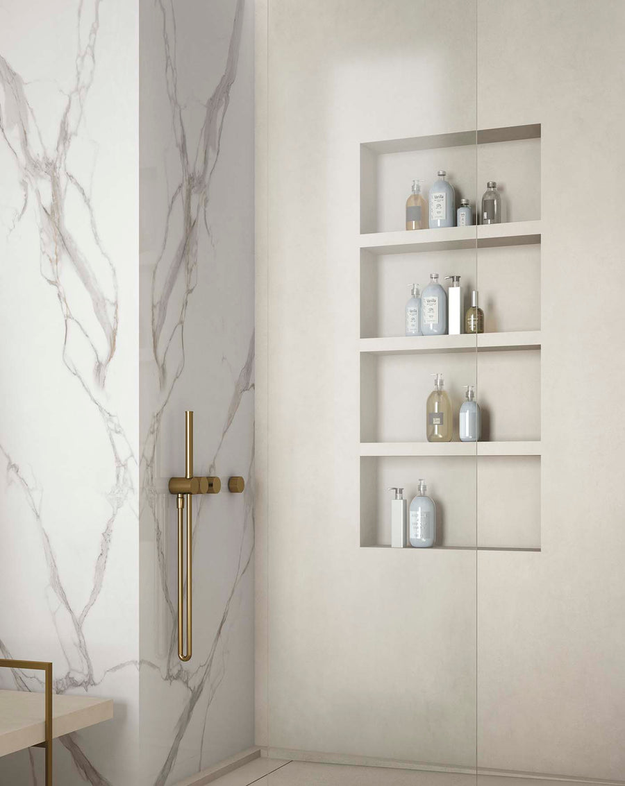 A modern shower with white marble walls, a gold shower fixture, and built-in shelves holding various bottles. The design is minimalist and elegant, with neutral tones and clean lines.