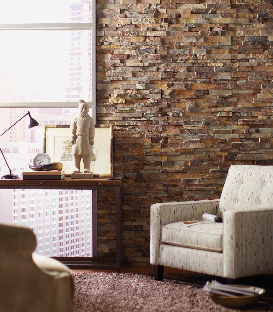 A cozy living room with a stone accent wall, armchairs, a brown rug, and a side table holding a lamp, clock, books, framed art, and a statue by a window with a city view.