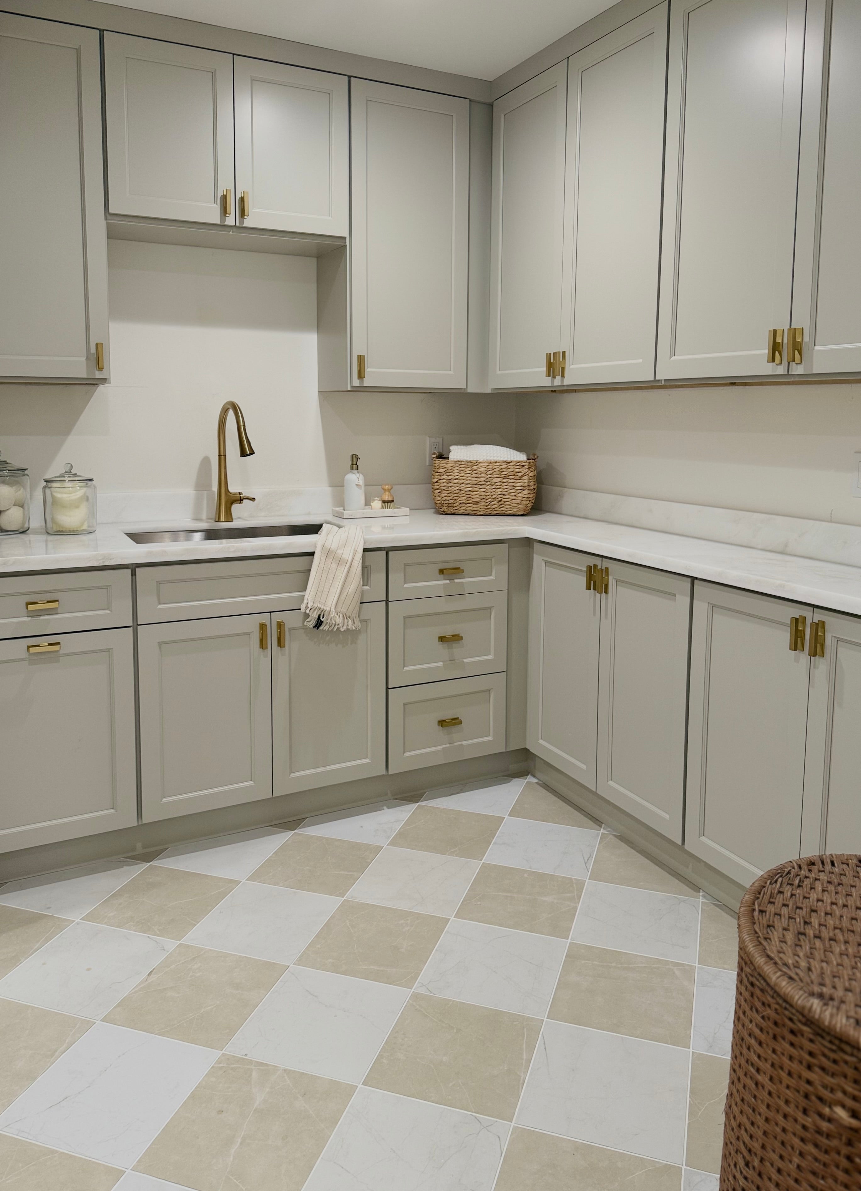 
		                            	A modern kitchen with light gray cabinets, gold hardware, white marble countertops, a gold faucet, and a checkerboard floor in beige and Snow Callie 12x12 Ceramic Tile. A basket, jars, and a towel are arranged near the sink.
		                            