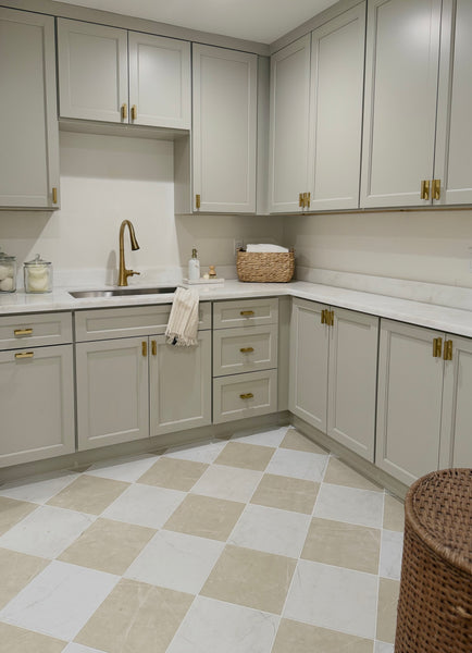 A modern kitchen with light gray cabinets, gold handles, a marble countertop, a gold faucet, a Callie 12x12 Ceramic Tile floor in Alabaster, a wicker basket, glass jars, and a towel hanging on the cabinet.