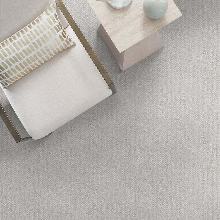 Top view of a modern, minimalist setting with a light-colored armchair, a geometric pillow, and a small square side table holding a round decorative object, all placed on a textured light gray carpet.